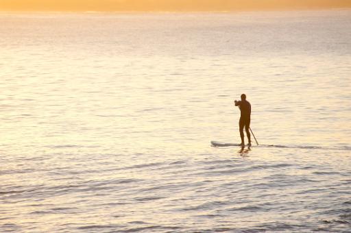 Man on a paddle board paddling across a calm ocean parallel to a sandy tropical beach, silhouetted against bright water with copyspace